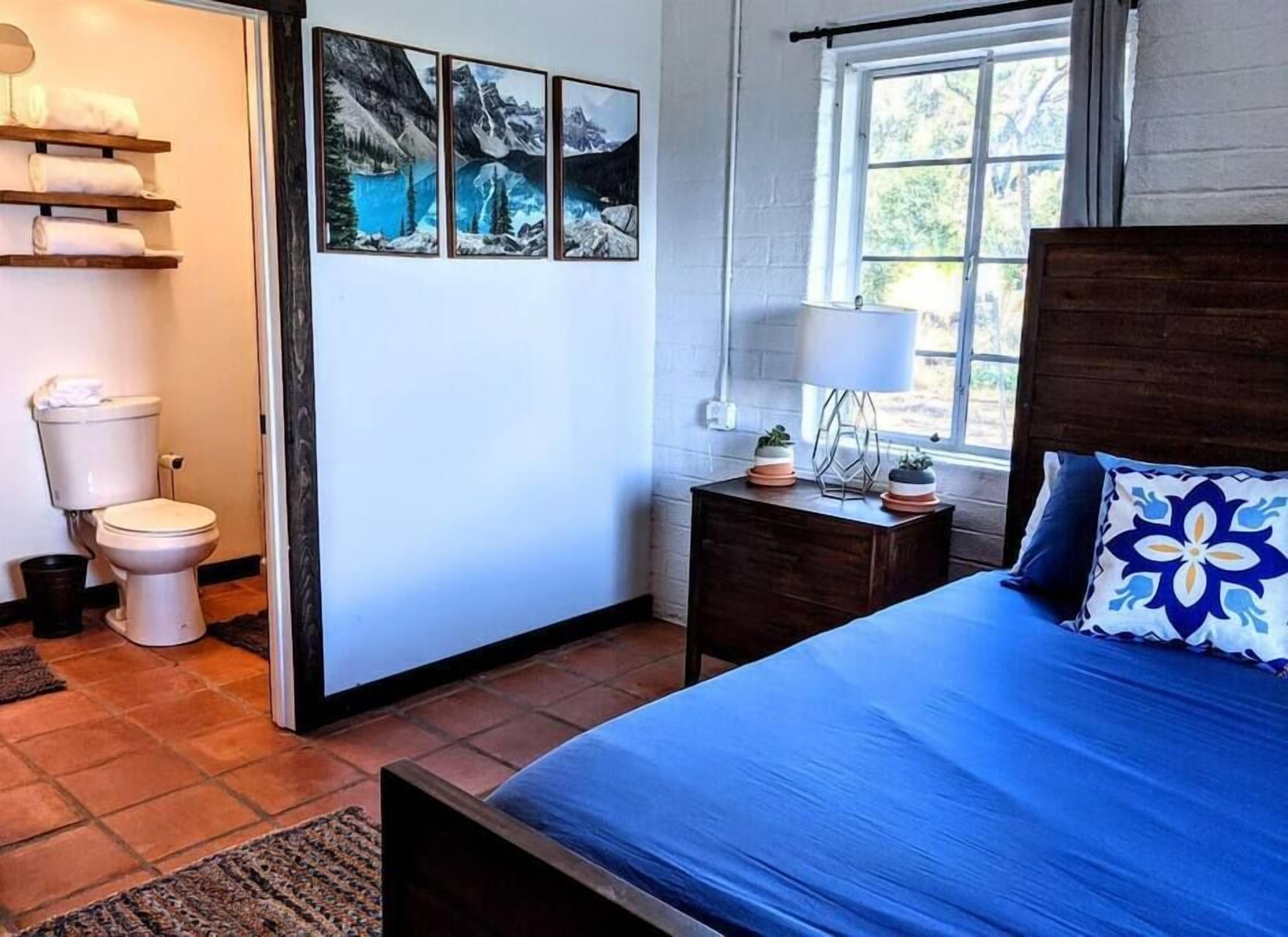 A bedroom with a bed, a nightstand with a lamp, a window, and a bathroom with a toilet and shelves.