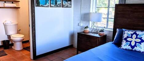 A bedroom with a bed, a nightstand with a lamp, a window, and a bathroom with a toilet and shelves.