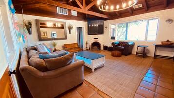 A spacious living room with a large sofa, a coffee table, and a fireplace. There is a television mounted on the wall and a window providing natural light.