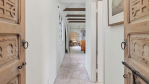 A narrow hallway with wooden doors on either side, a wicker chair in the center, and a bed with a brown blanket in the background. - Casa Blanca, Secluded Mediterranean Retreat in RSF (Rancho Santa Fe)