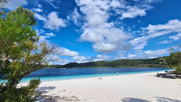 Una playa con aguas azules cristalinas, arena blanca y una colina boscosa al fondo.