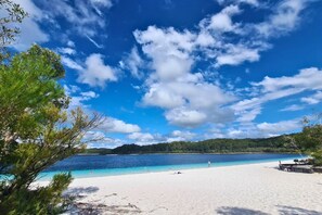 Una playa con aguas azules cristalinas, arena blanca y una colina boscosa al fondo.