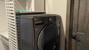 A compact, modern laundry room with a built-in washer and dryer, a white shelving unit above, and a wooden door to the right.