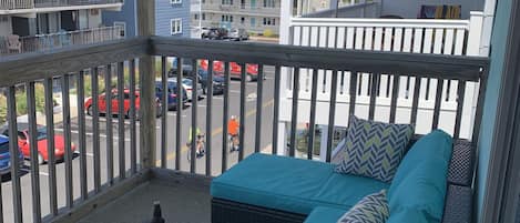A balcony with wicker furniture and a table, overlooking a street with parked cars and buildings.