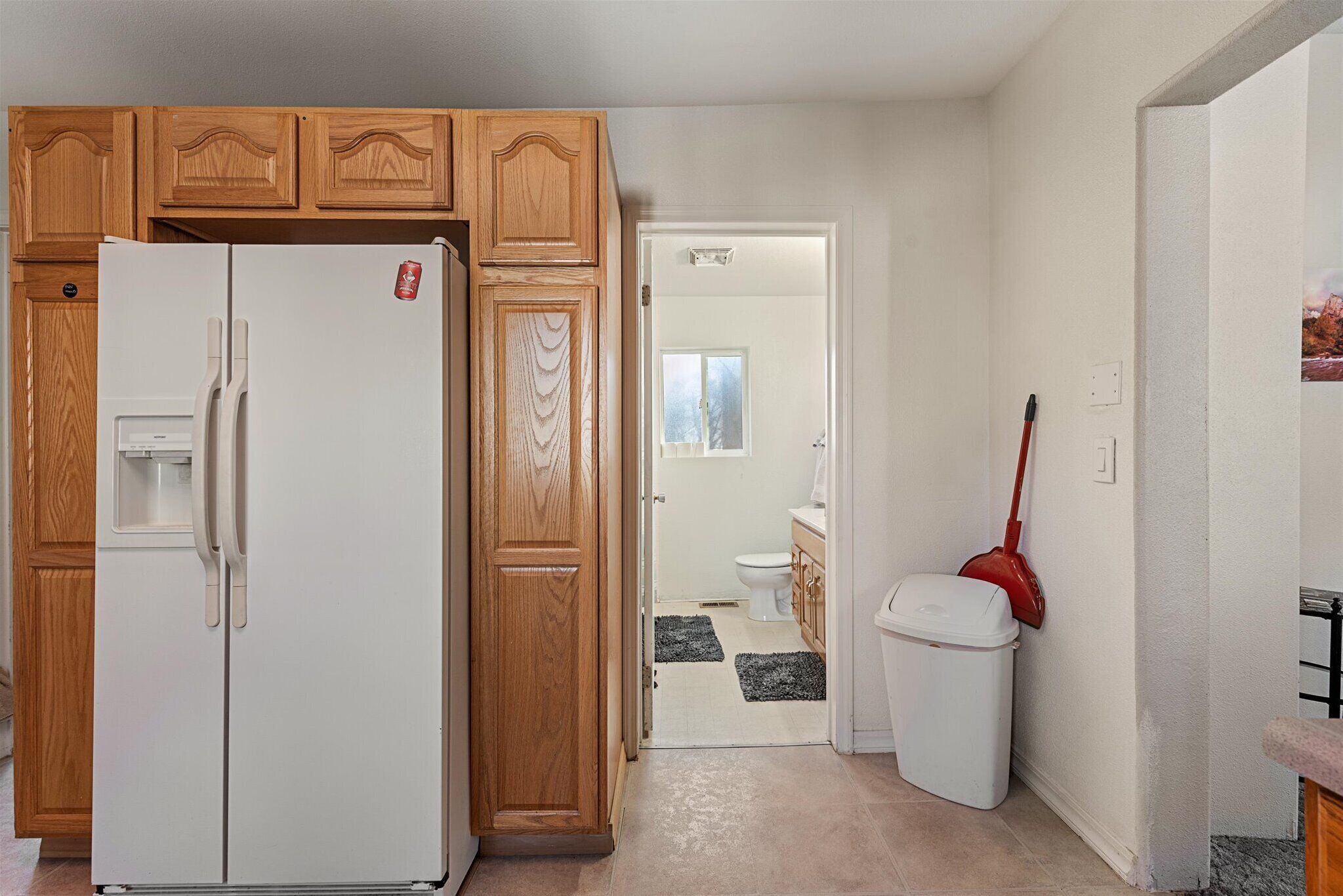 A kitchen with wooden cabinets, a white refrigerator, a white trash bin, and a red mop leaning against the wall.