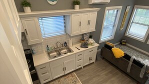 Une cuisine moderne avec des meubles blancs, un évier et un plan de travail. On y trouve un canapé gris avec un coussin jaune et une petite table sur laquelle est posée une plante.