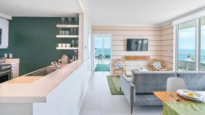 Una cocina moderna con encimera blanca, horno empotrado y fregadero. Hay una zona de comedor con un sofá gris, una mesa de madera y un televisor montado en la pared.