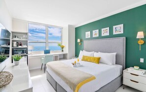 A modern hotel room with a large bed, a desk with a laptop, a view of the sea, and artwork on the walls.