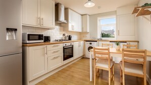 A modern kitchen with white cabinets, a built-in oven, a microwave, and a washing machine. There is a wooden dining table with chairs and a pendant light above. - Shore View - Donnini Apartments (Prestwick)