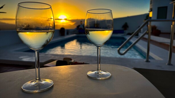 Twee glazen witte wijn op een tafel, met een zwembad en een zonsondergang op de achtergrond.