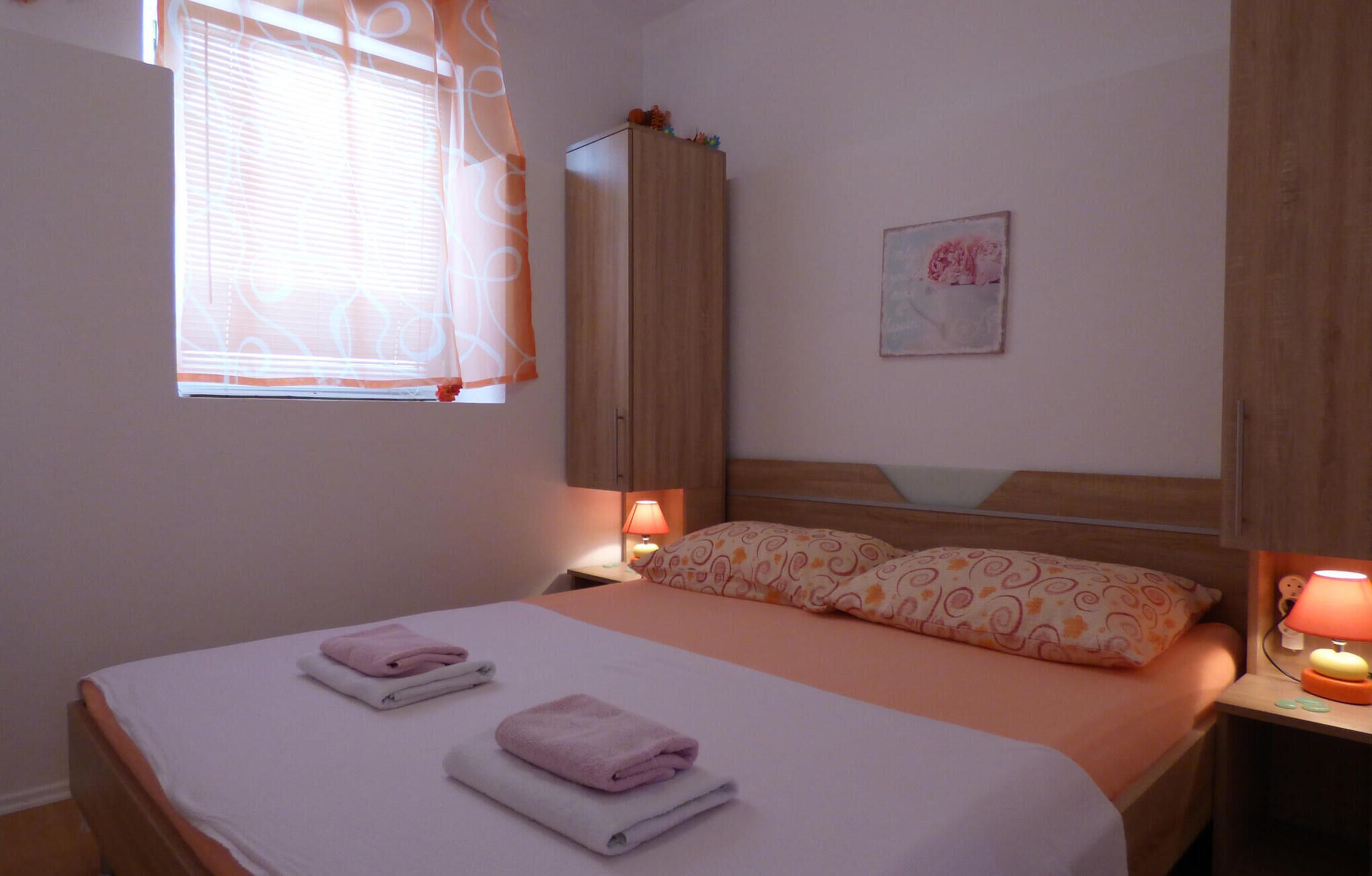 A bedroom with a bed, two bedside lamps, a window with blinds, a wooden wardrobe, and a wall-mounted painting.
