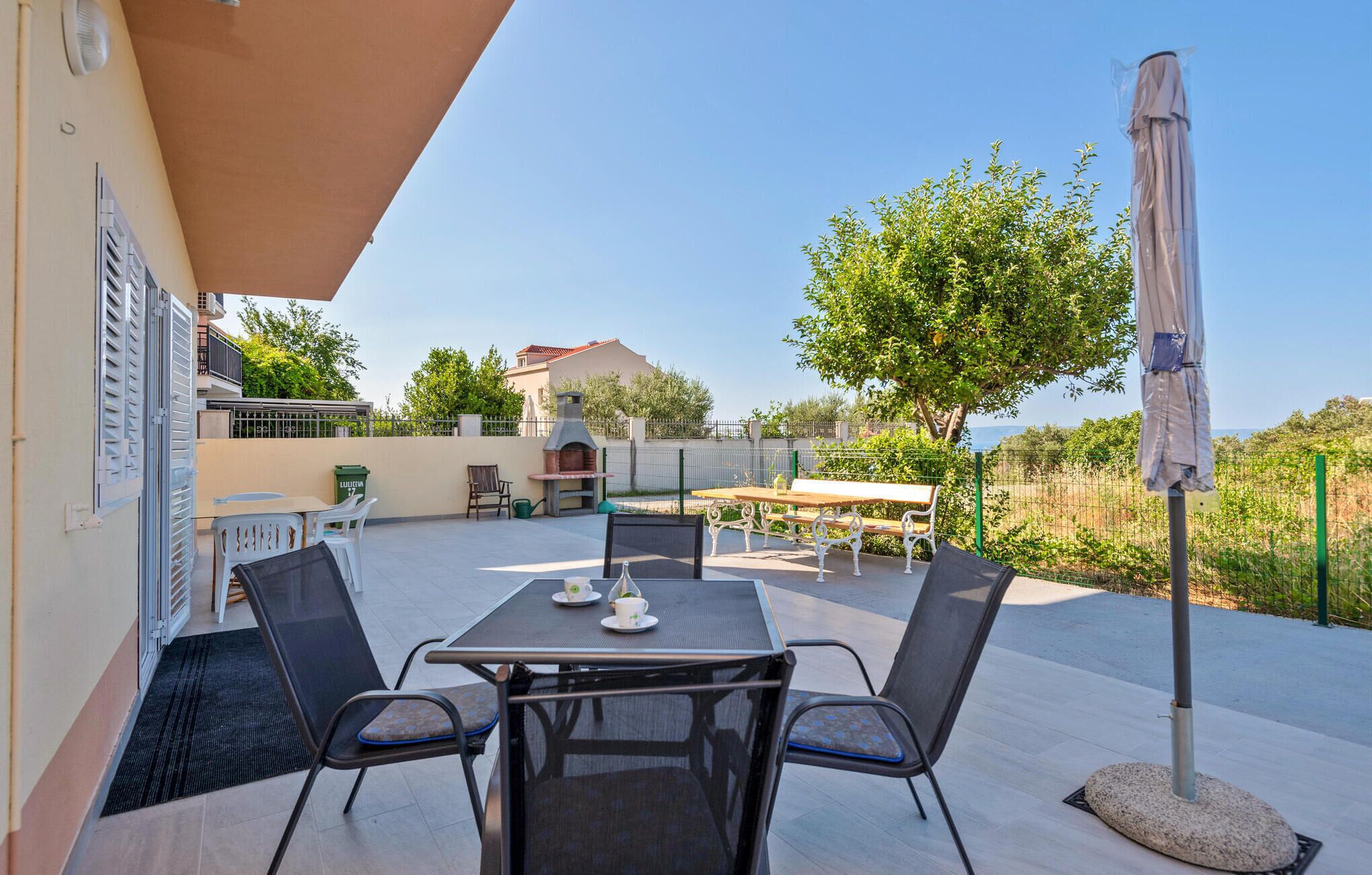 Eine Terrasse im Freien mit Tisch und Stühlen, einem Grill und Blick auf einen Baum und ein Gebäude im Hintergrund.