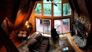 A cozy loft-style room with a stone fireplace, a large window overlooking a deck, and a comfortable seating area with a sofa and a coffee table. - HOT TUB,  Unique A-Frame Cabin, Walk to ski resort (Mars Hill)