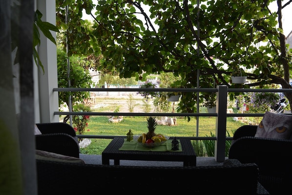 Un balcón con una mesa que sostiene fruta y un farol, con vista a un jardín y un edificio.