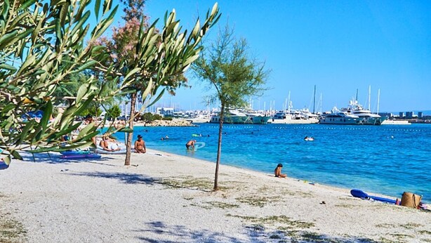 Une plage avec des bateaux en arrière-plan, un ciel dégagé et des gens se détendant au bord de l’eau.