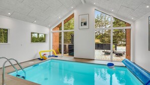 A modern indoor pool with clear glass walls, a wooden deck, and a view of the surrounding trees. - Lovely home in Blåvand with sauna (Blåvand)