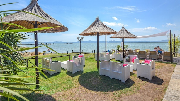 An outdoor seating area with wicker chairs and thatched umbrellas, overlooking a body of water with a distant view of mountains.