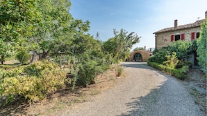 A gravel path leading to a stone building with red shutters, surrounded by lush greenery and a clear sky.