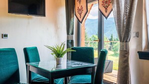 Habitación con mesa de vidrio, dos sillas verdes, una planta en maceta, un televisor de pantalla plana y vista a las montañas a través de una ventana con cortinas decorativas.