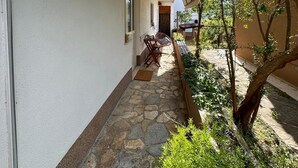 Un étroit sentier pavé de pierres mène à un petit espace extérieur aménagé avec une table et des chaises, le tout entouré de verdure et d’un bâtiment doté d’une porte en bois.