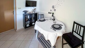 Un coin repas avec une table ronde dressée pour deux personnes, une chaise noire, un téléviseur fixé au mur, une étagère avec des objets décoratifs et une porte blanche.