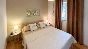 Un lit bien fait, avec une couette blanche et des oreillers, dont l’un est brodé du mot « JOIE ». On y trouve une petite table de chevet avec une lampe, une œuvre d’art accrochée au mur et une fenêtre avec des rideaux.