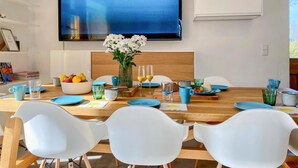 Un comedor con una mesa de madera, sillas blancas, un florero con flores y un televisor de pantalla plana grande en la pared.