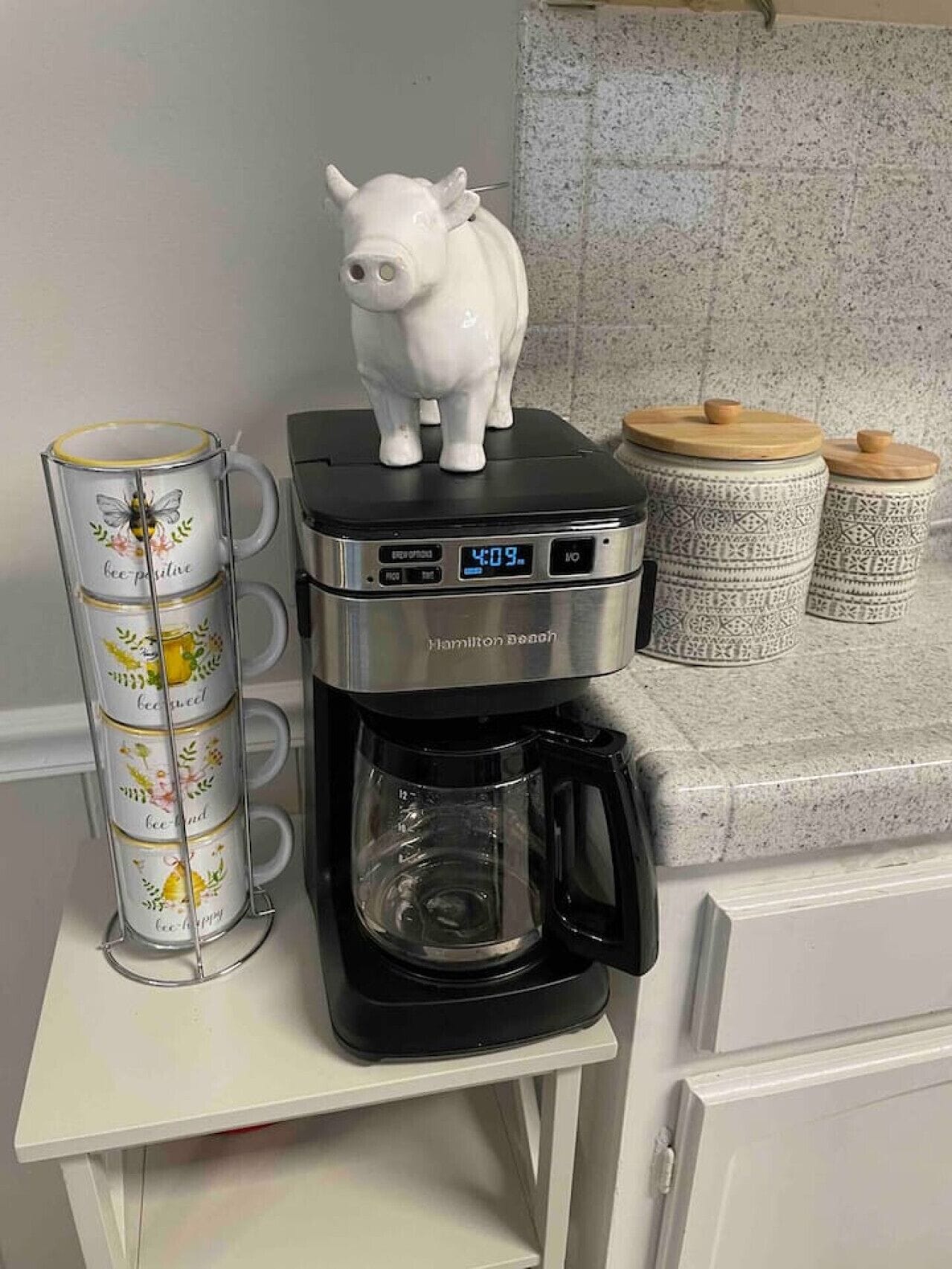 A Hamilton Beach coffee maker on a white table with a pig figurine and a mug rack with bees and flowers.