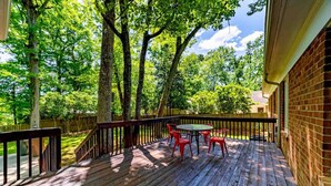 A wooden deck with a brick wall, surrounded by trees, and a table with chairs. - Modern 4 Bedroom pets welcome sleeps 8 (Greensboro)