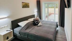 Una cama bien hecha con un edredón gris y una almohada con forma de corazón, una mesita de noche con un control remoto, y vistas al exterior a través de una puerta de cristal.