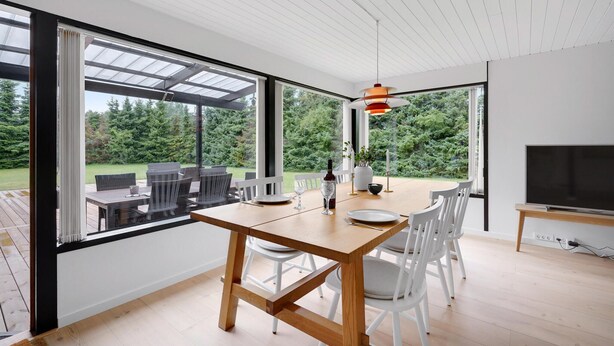 A modern dining area with a wooden table and white chairs, large windows offering a view of a garden, and a flat-screen TV on the wall. - 4 bedroom amazing home in Væggerløse (Væggerløse)