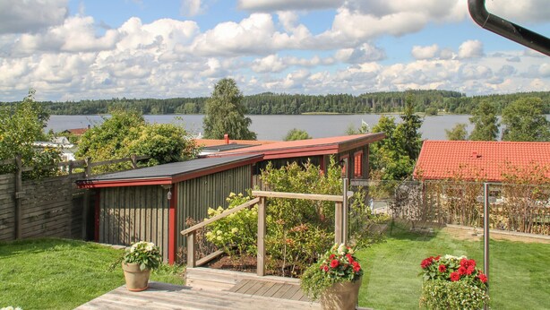 A wooden deck with a red roof, a small garden with potted plants, and a view of a lake and forest in the background.