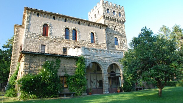 Un edificio histórico de piedra con una entrada central en arco, una torre y un jardín bien cuidado.