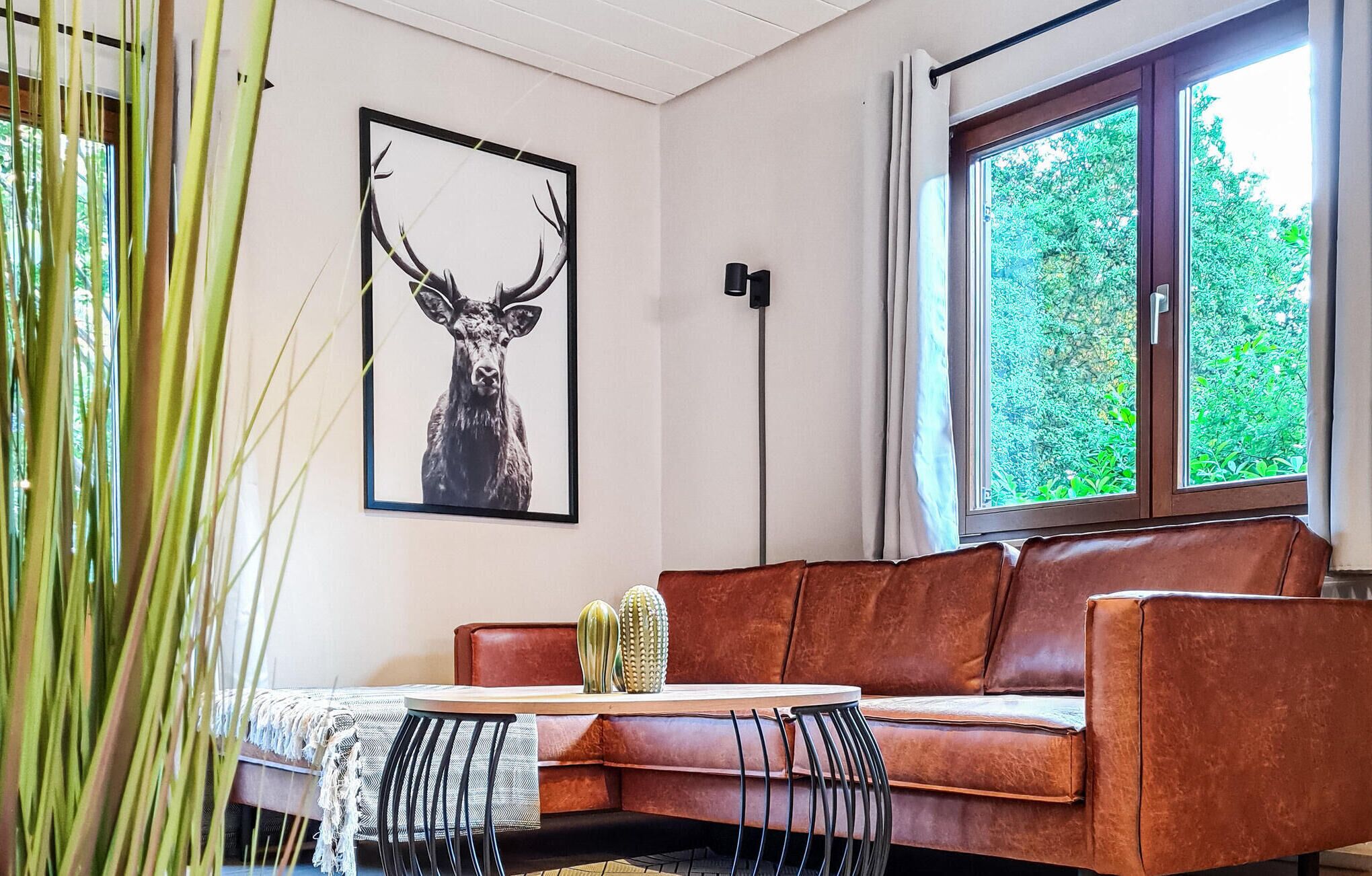 A living room with a brown leather sofa, a cactus, a framed deer antler artwork, a tall floor lamp, and a window with a view of greenery.