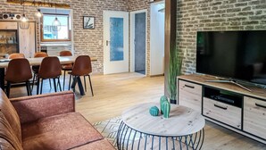 A modern living room with a brick wall, a TV, a dining area with chairs, and a kitchen in the background. - Ferienhaus 16 in Kirchheim (Kirchheim)