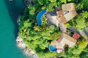 Vista aérea de una propiedad costera con piscina, varias construcciones y abundante vegetación.