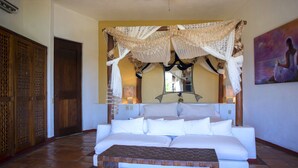 A room with a white sofa, a wooden coffee table, a decorative canopy, a wooden door, and a painting on the wall. - The Estate at Pacific Vacation | StayPV Luxury Oceanfront with Staff (Zihuatanejo)