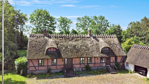 Ett traditionellt hus med halmtak och en grund av sten, en trätrappa som leder upp till entrén och en grusuppfart.