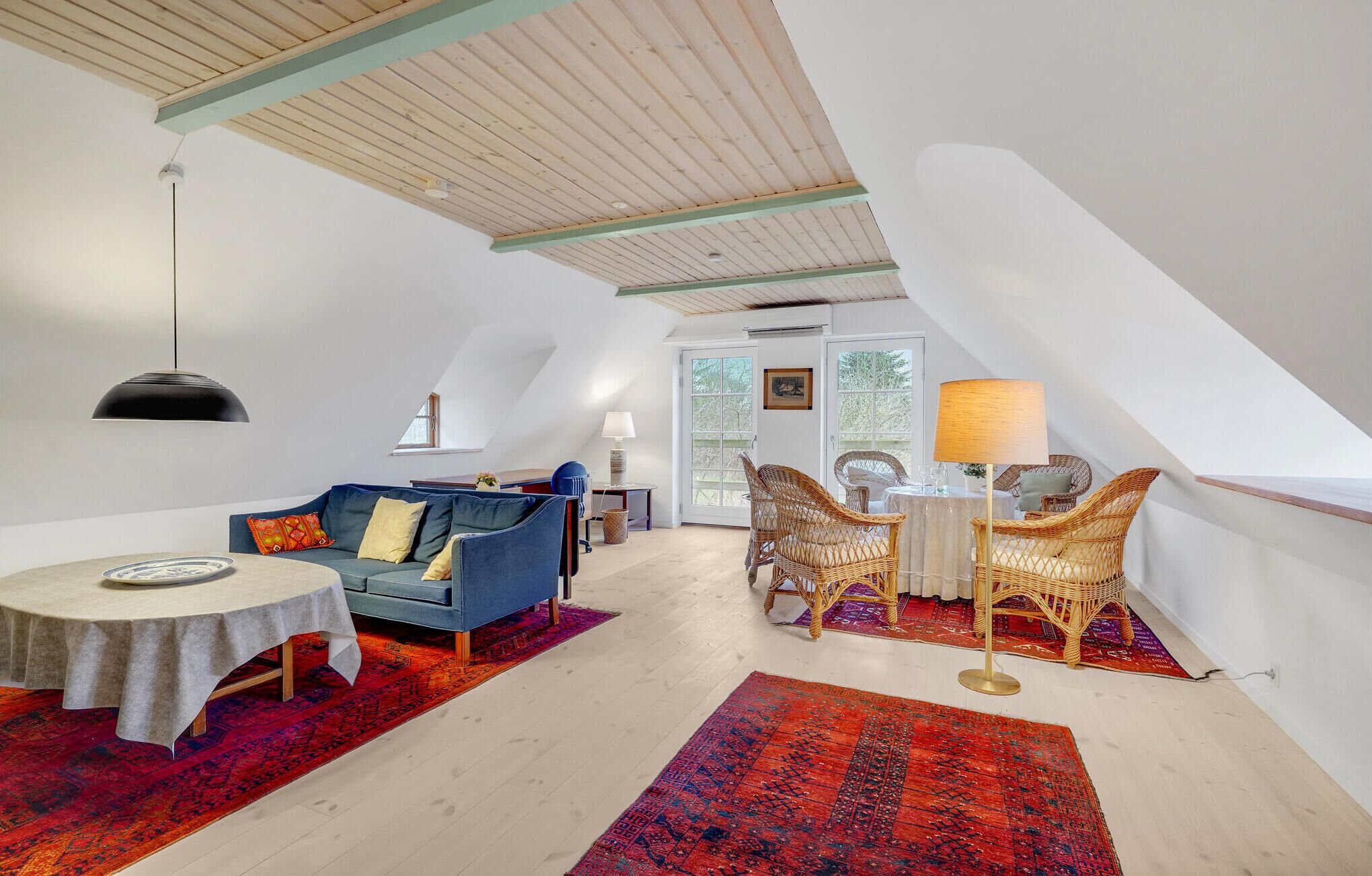 A cozy loft-style room with a high ceiling, a blue sofa, wicker chairs, a small dining table, and a red patterned rug.