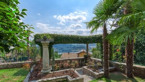 A garden with a stone pillar, a pergola covered in greenery, and a view of a distant town.