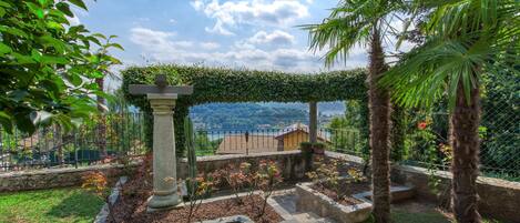 A garden with a stone pillar, a pergola covered in greenery, and a view of a distant town.