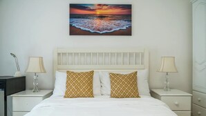 A hotel room with a bed, two bedside tables with lamps, a nightstand, and a wall-mounted painting of a sunset over the ocean. - Deluxe 2Bed Duplex w/parking (Greater London)