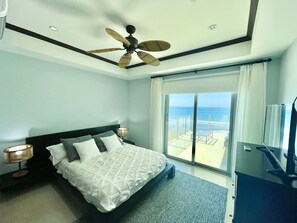 A hotel room with a bed, a ceiling fan, a large window with a view of the beach, and a TV.