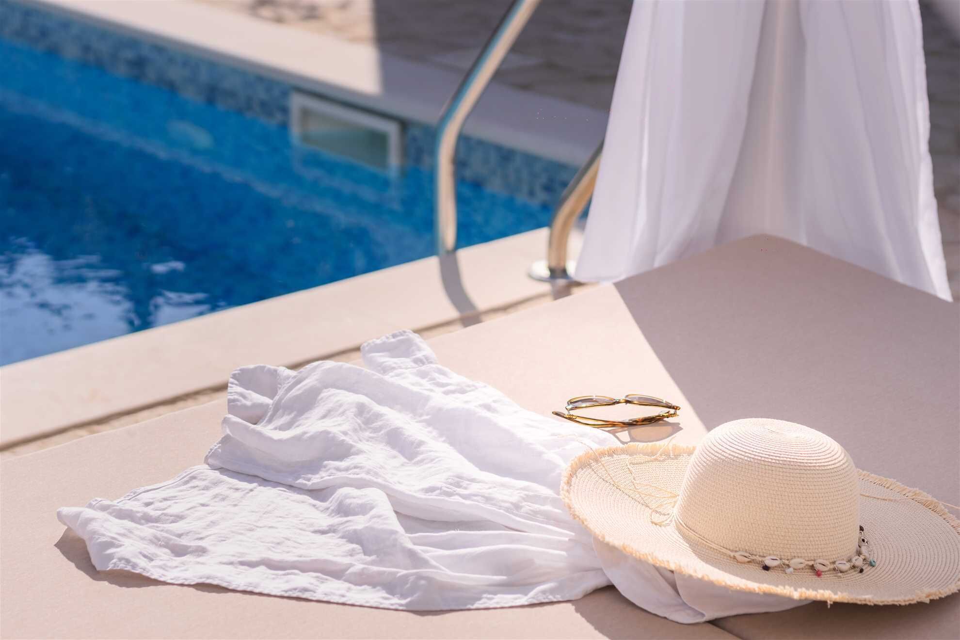 Ein Poolbereich mit weißem Handtuch, einem Strohhut und einer Sonnenbrille auf einer Liege.