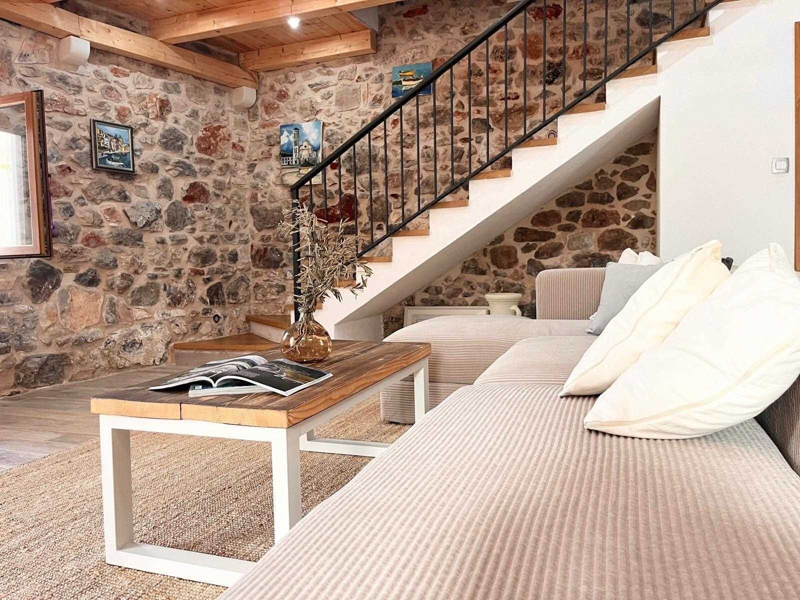A cozy living room with a stone wall, a wooden staircase, a sofa, and a coffee table with books.