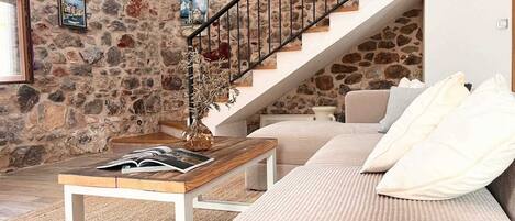 A cozy living room with a stone wall, a wooden staircase, a sofa, and a coffee table with books.