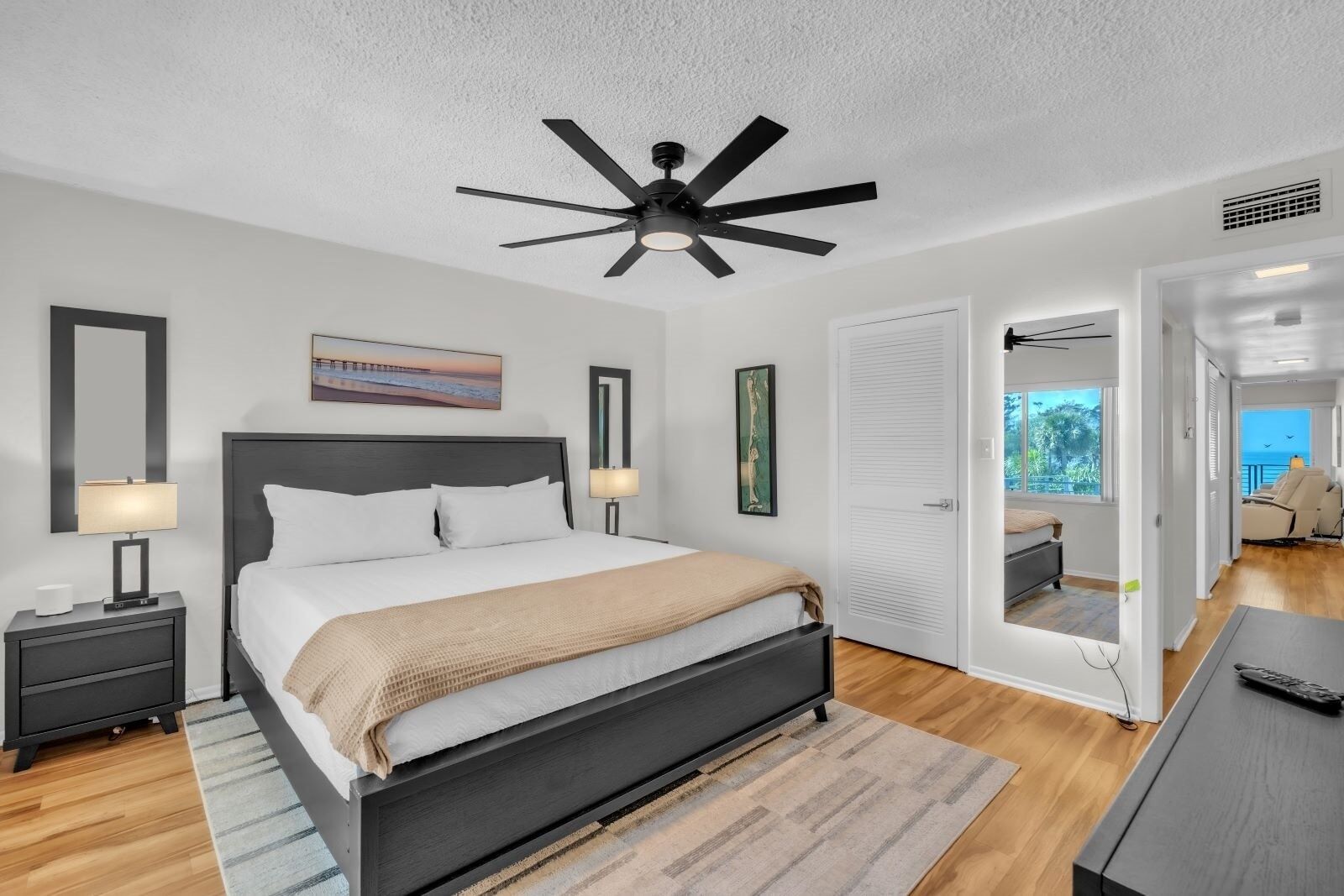 A modern bedroom with a bed, bedside tables, a ceiling fan, artwork, and a view of the outdoors.