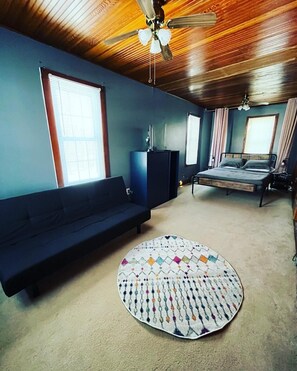 Une pièce avec un plafond en bois, un canapé bleu foncé, un lit avec une tête de lit, une petite table et un tapis à motifs.