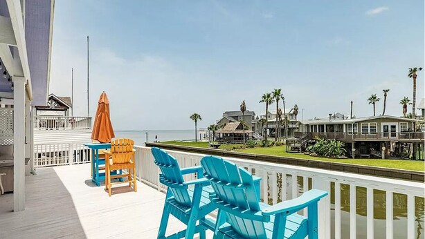 A deck with blue and orange Adirondack chairs, a small table, and an umbrella. The deck overlooks a body of water and a row of houses with palm trees.