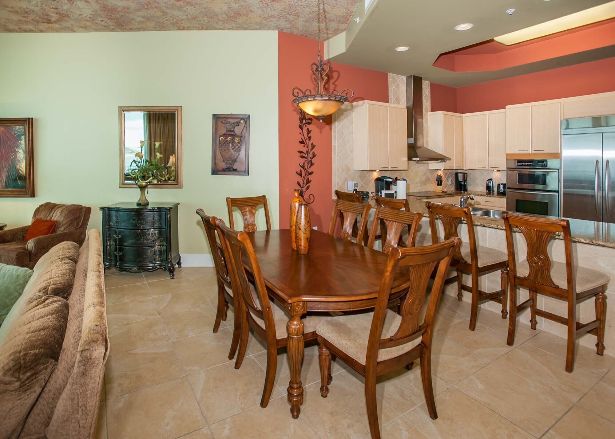 A dining area with a wooden table and chairs, a chandelier, and a kitchen with modern appliances.
